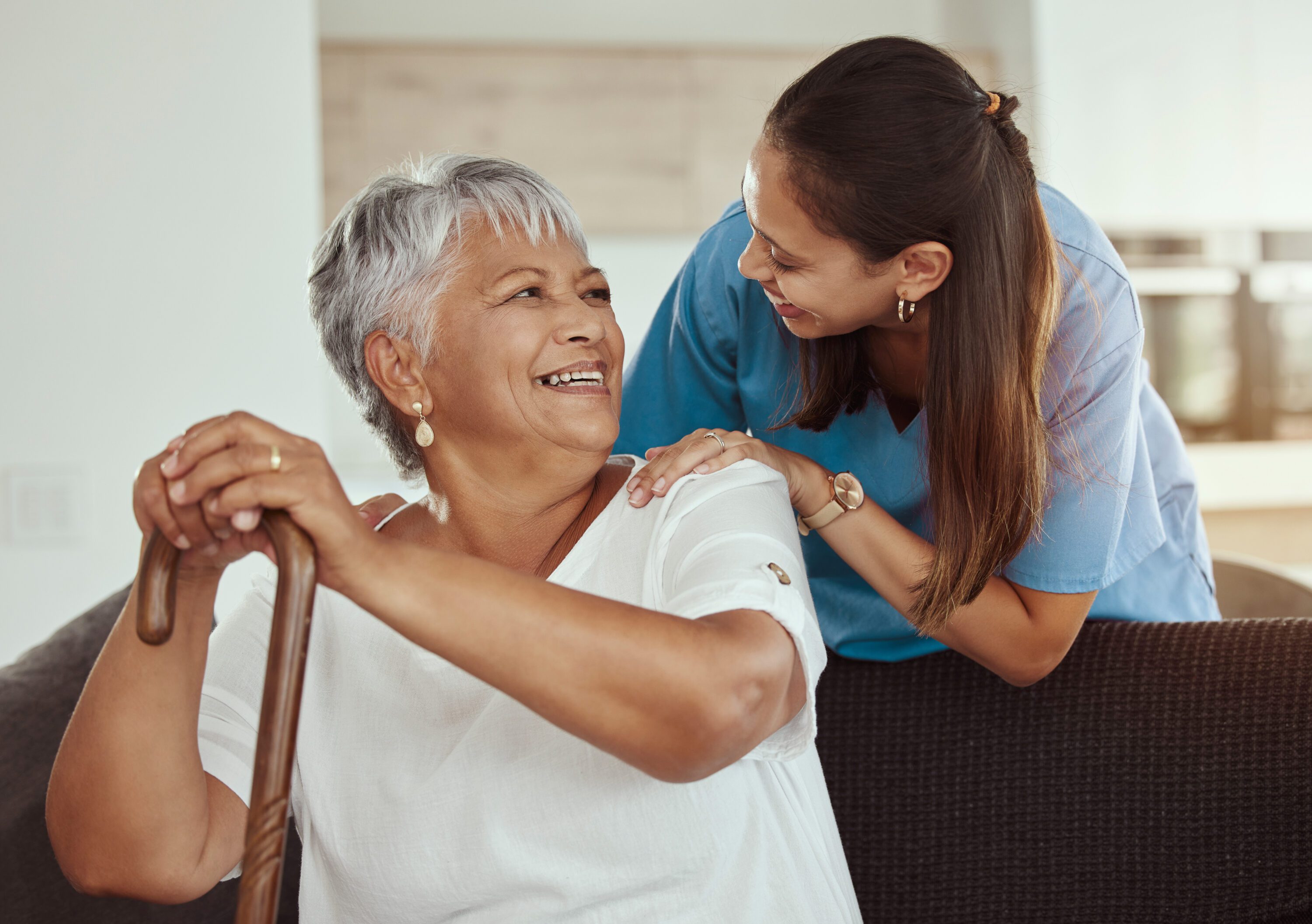 Happy, relax and senior woman with caregiver smile while sitting on a living room sofa in a nursing home. Support, help and professional nurse or healthcare worker helping elderly lady or patient.