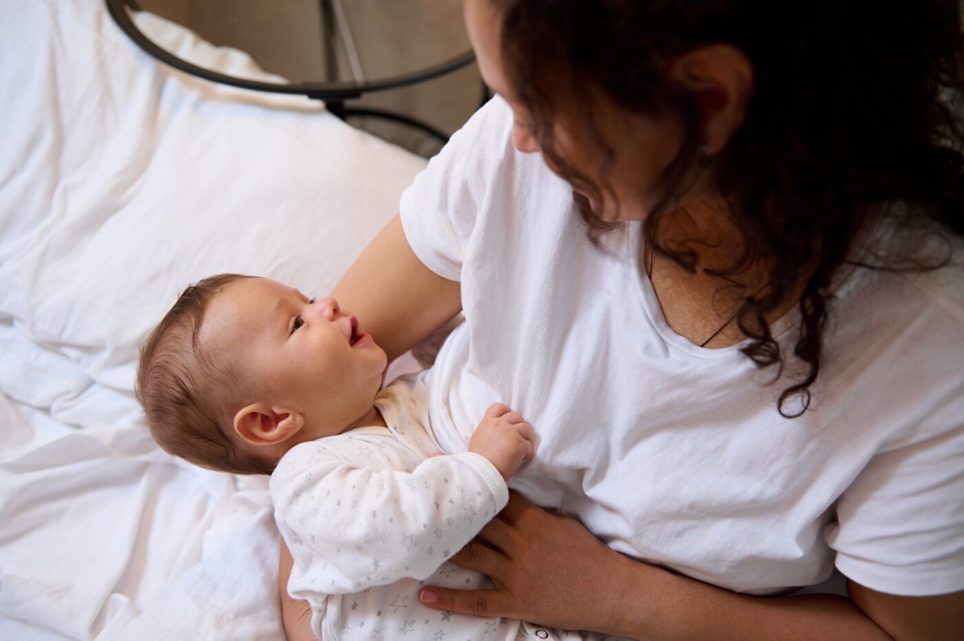 Authentic portrait of curly brunette young adult woman, happy loving mother smiling, expressing positive emotions while breastfeeding and cuddling her newborn baby.