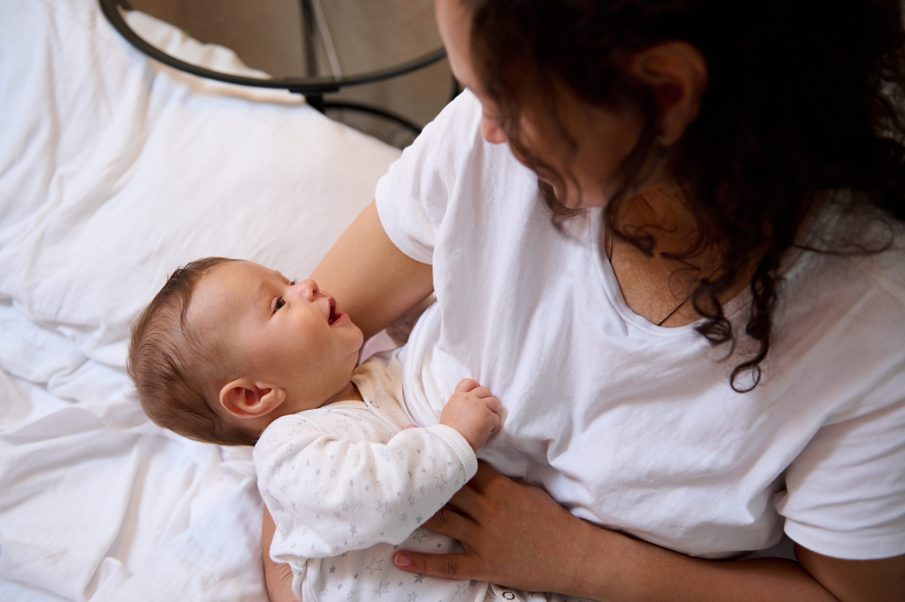 Authentic portrait of curly brunette young adult woman, happy loving mother smiling, expressing positive emotions while breastfeeding and cuddling her newborn baby.