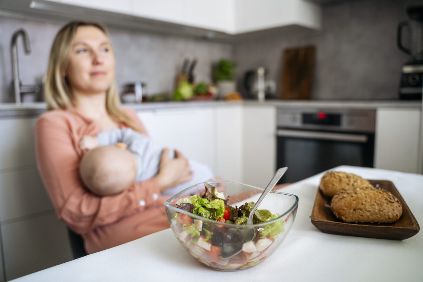 Fresh food on dining table with mother breastfeeding toddler son in kitchen.