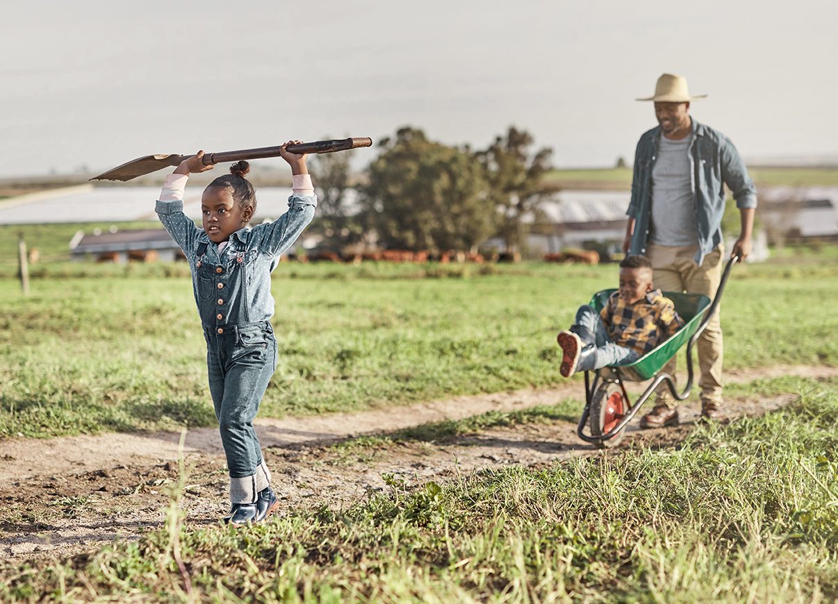 A father and his two children, one boy and one girl, walking on a farm road with cows in the distance.