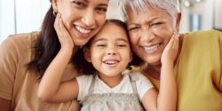 Three generations of women—a grandmother, mother, and young child—smiling closely together with their heads touching and hands resting on their cheeks in a bright indoor setting.