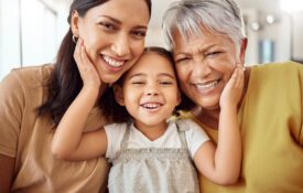 Three generations of women—a grandmother, mother, and young child—smiling closely together with their heads touching and hands resting on their cheeks in a bright indoor setting.