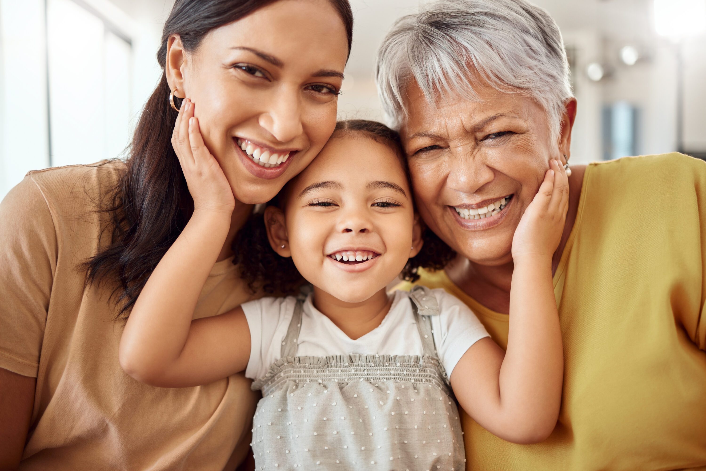 Three generations of women—a grandmother, mother, and young child—smiling closely together with their heads touching and hands resting on their cheeks in a bright indoor setting.