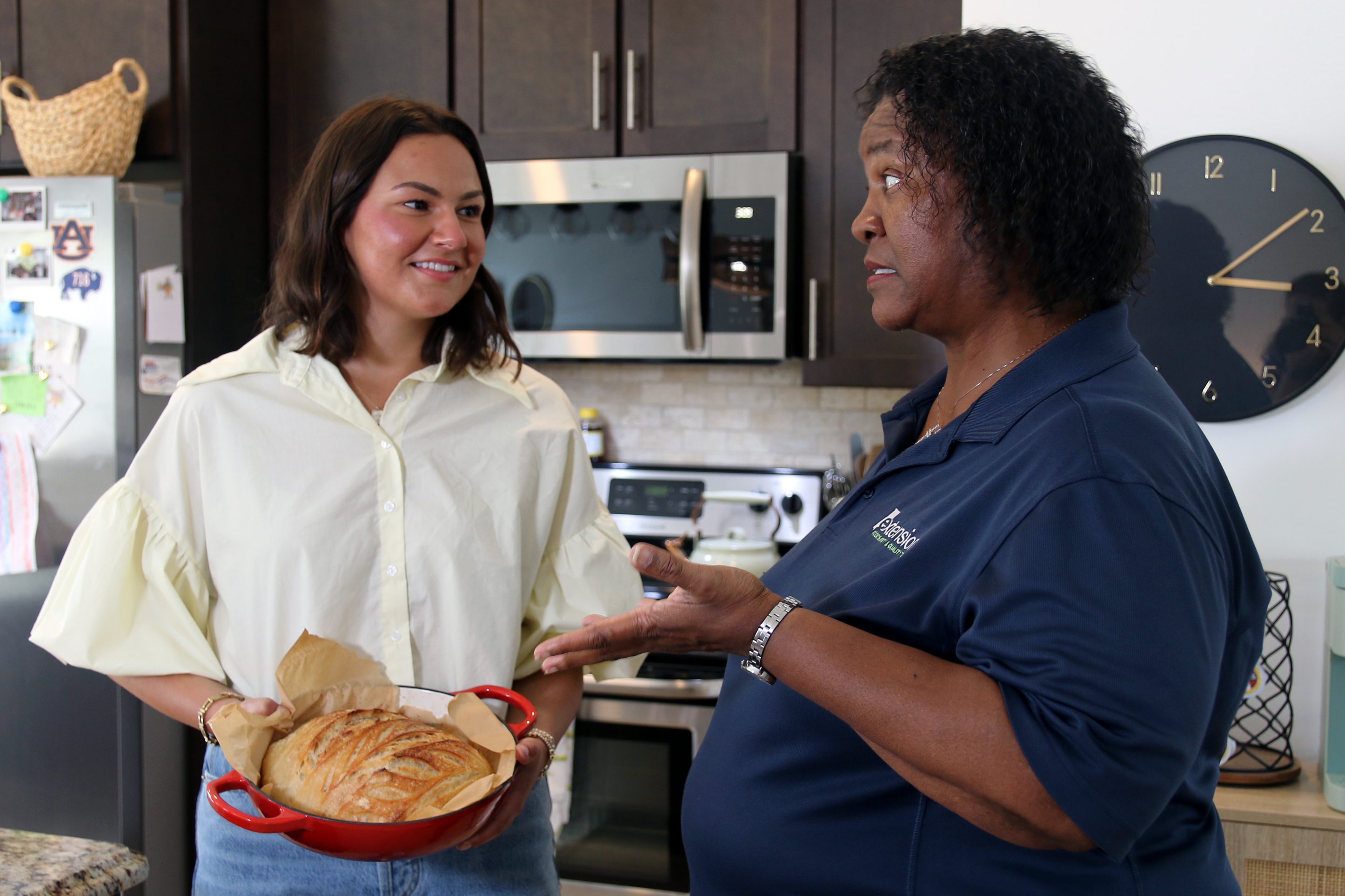 Elizabeth Belcher and Janet Johnson talking in Elizabeth's kitchen while she holds a pan of sourdough bread.