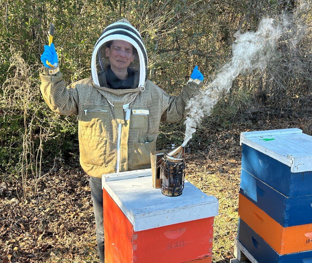 A frustrated beekeeper in protective gear near a hive with a smoker.