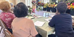 Jim DelPrince works on a floral arrangement with a spread of flowers to his side. In the foreground, three people are sitting and watching Jim's presentation.