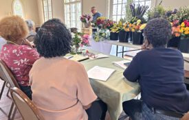 Jim DelPrince works on a floral arrangement with a spread of flowers to his side. In the foreground, three people are sitting and watching Jim's presentation.