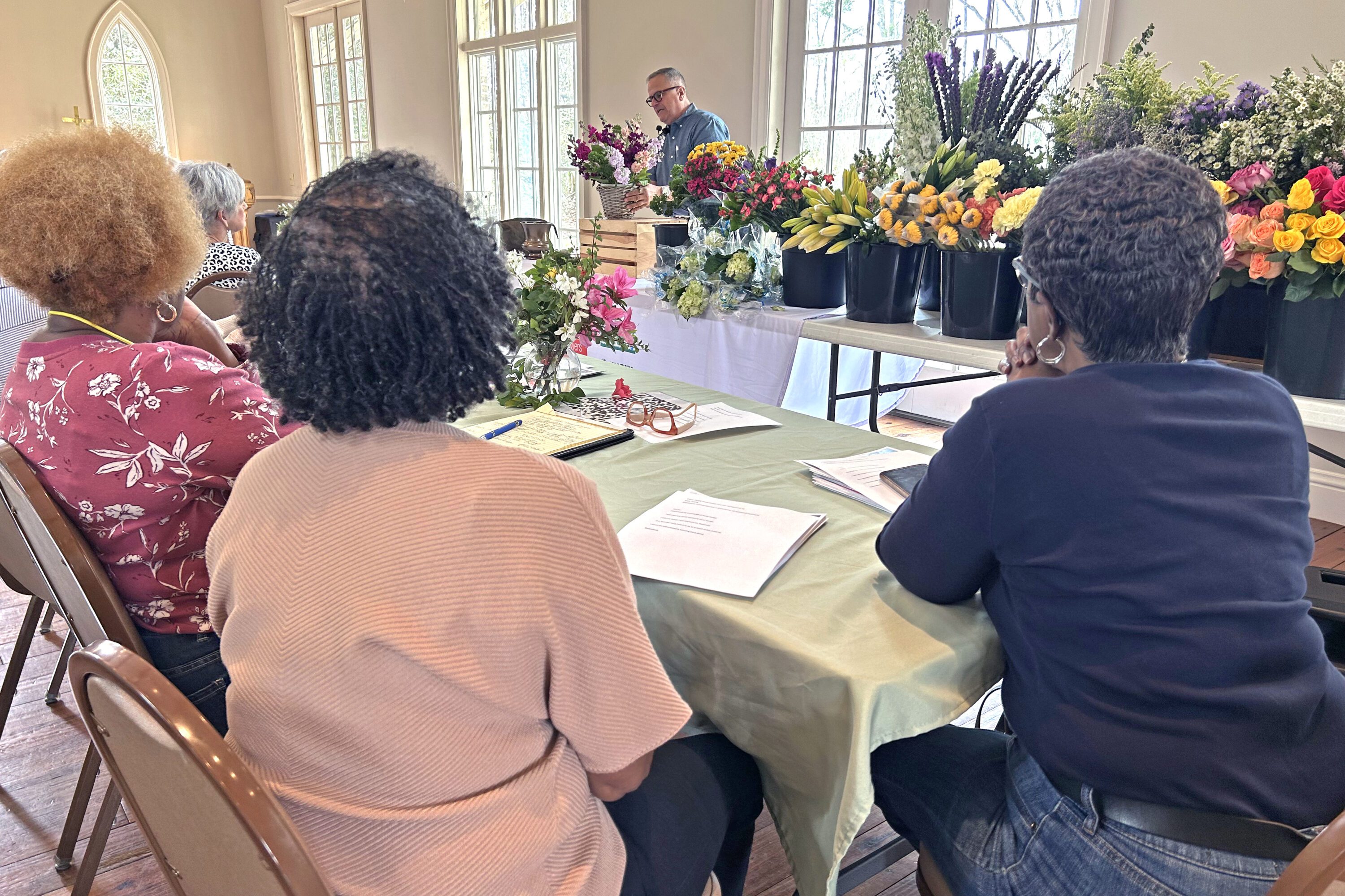 Jim DelPrince works on a floral arrangement with a spread of flowers to his side. In the foreground, three people are sitting and watching Jim's presentation.