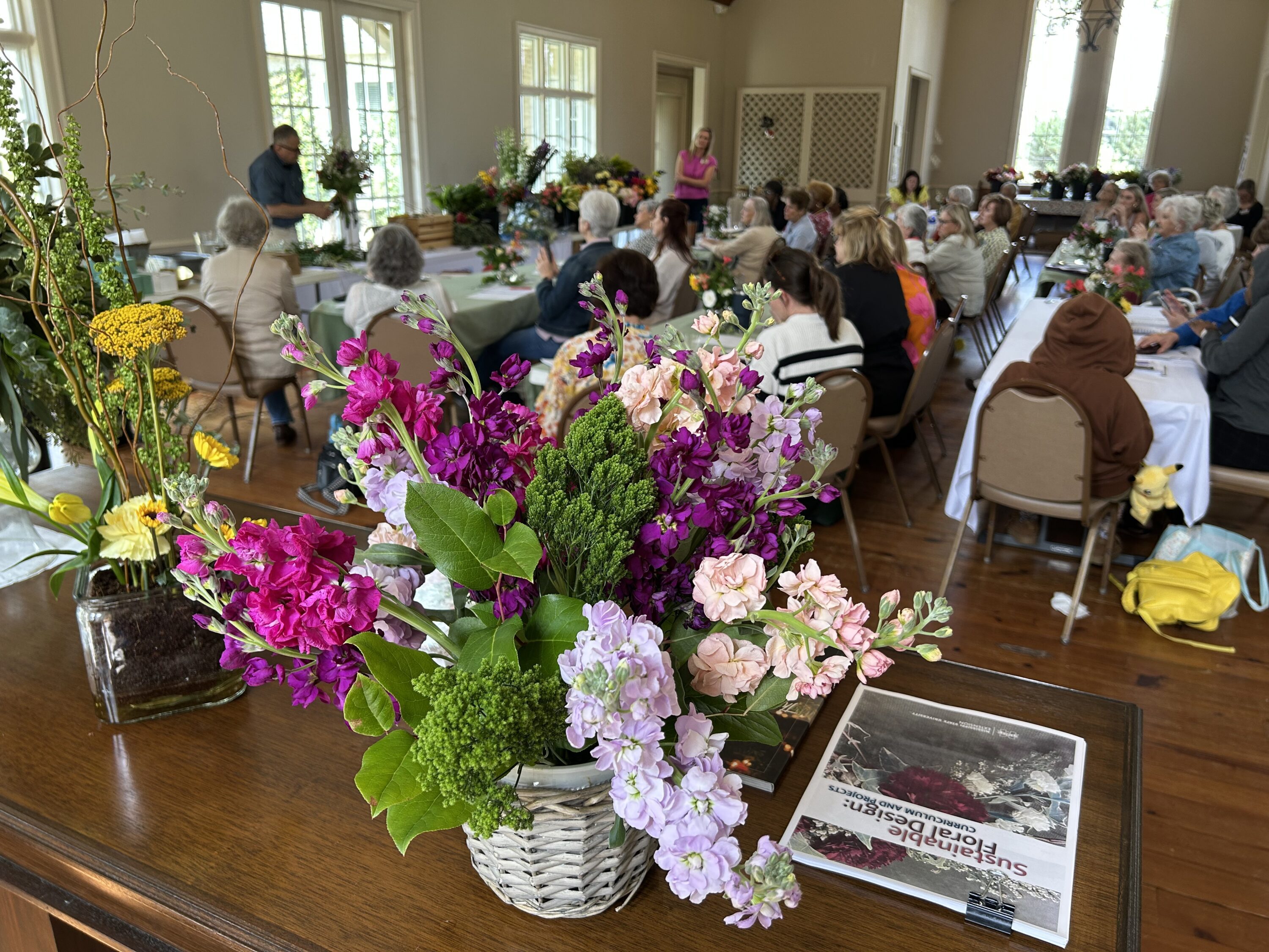A finished floral arrangement of purple and pinks in the foreground with a group of people sitting, watching a demonstration in the background.