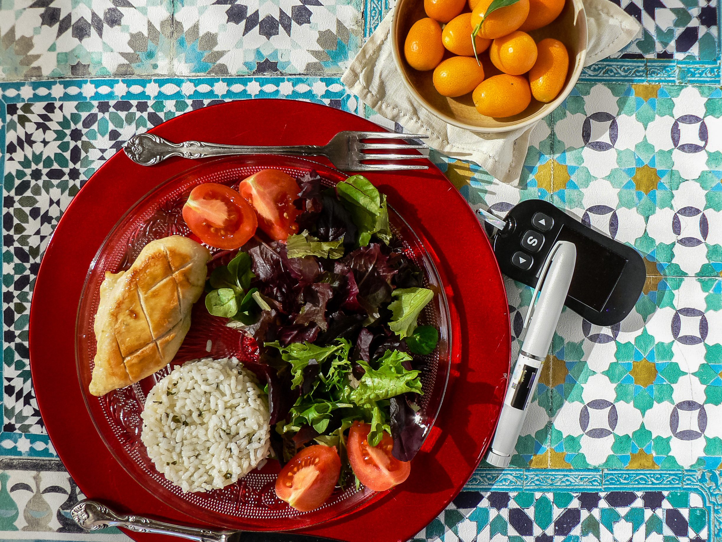 A red plate filled with vegetables, a lean protein, and rice. Also, a small bowl of citrus and a diabetes monitor.