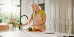Senior woman wearing apron, standing at a kitchen sink with water running and vegetables on a cutting board. The kitchen is very clean.
