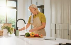 Senior woman wearing apron, standing at a kitchen sink with water running and vegetables on a cutting board. The kitchen is very clean.