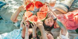 Family lying on picnic blanket and holding up pieces of watermelon for a fun photo - an example of keeping health simple.