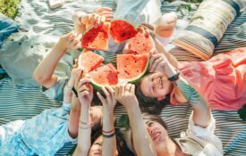 Family lying on picnic blanket and holding up pieces of watermelon for a fun photo - an example of keeping health simple.