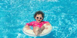 Little girl with pink glasses, curly hair and fuchsia swimsuit, enjoys the blue water pool with her round float on a sunny day