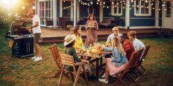 A group gathered around a table outside with a man standing near a grill and a woman bringing a salad toward the table.