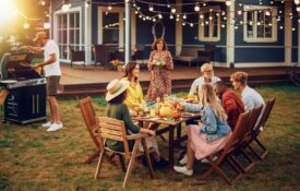 A group gathered around a table outside with a man standing near a grill and a woman bringing a salad toward the table.