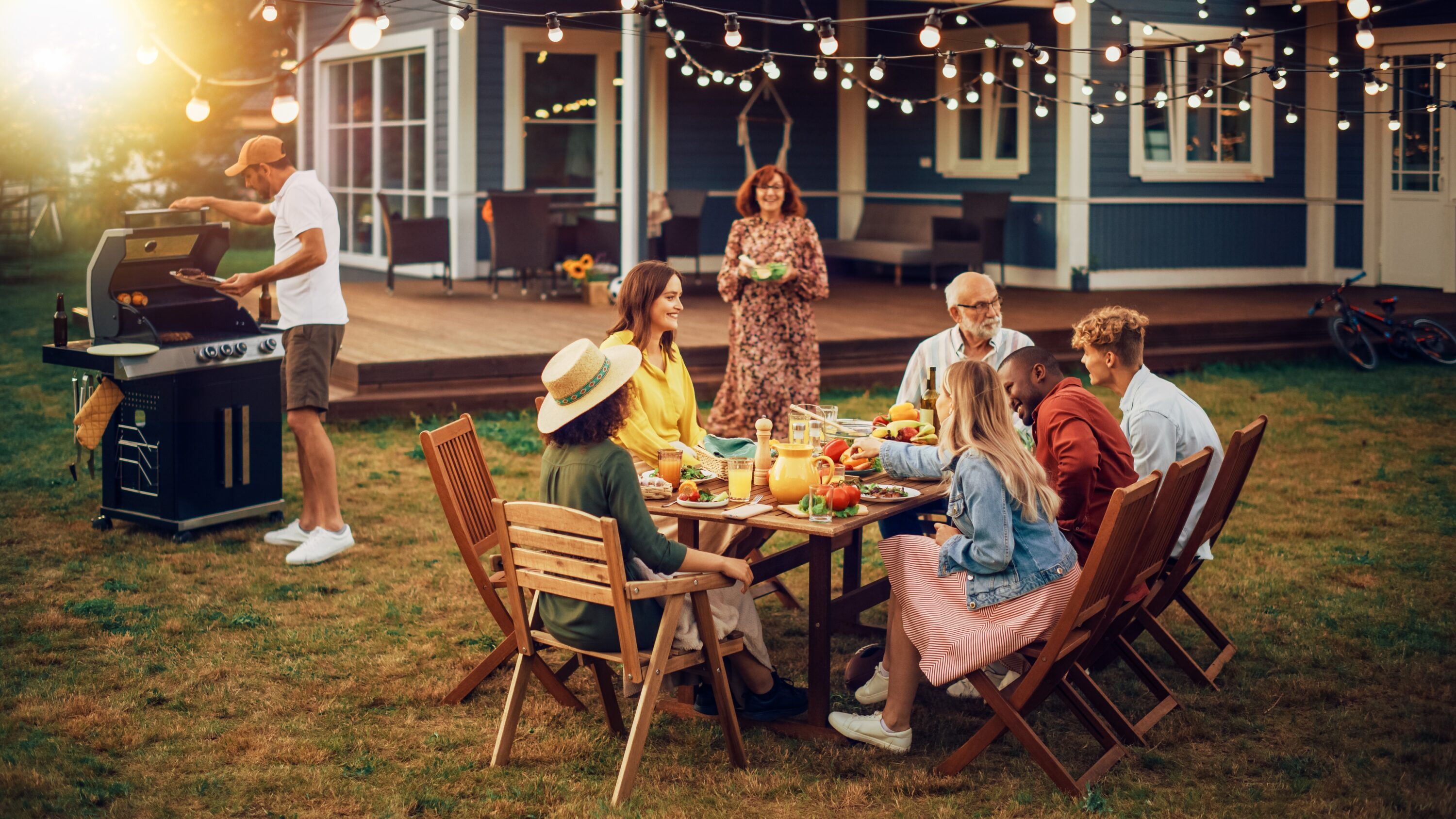 A group gathered around a table outside with a man standing near a grill and a woman bringing a salad toward the table.