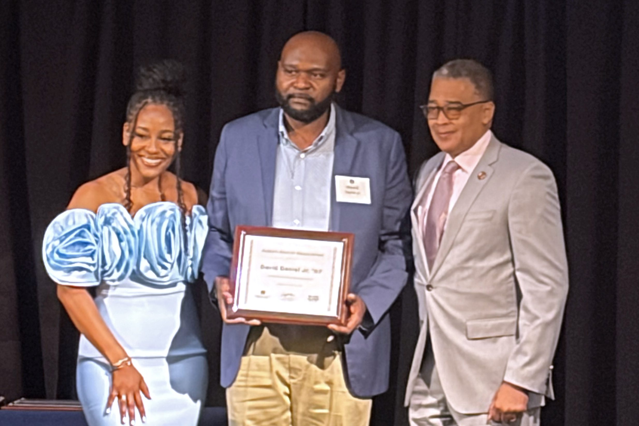 Three people standing on a stage. David Daniel is in the center, accepting his award.