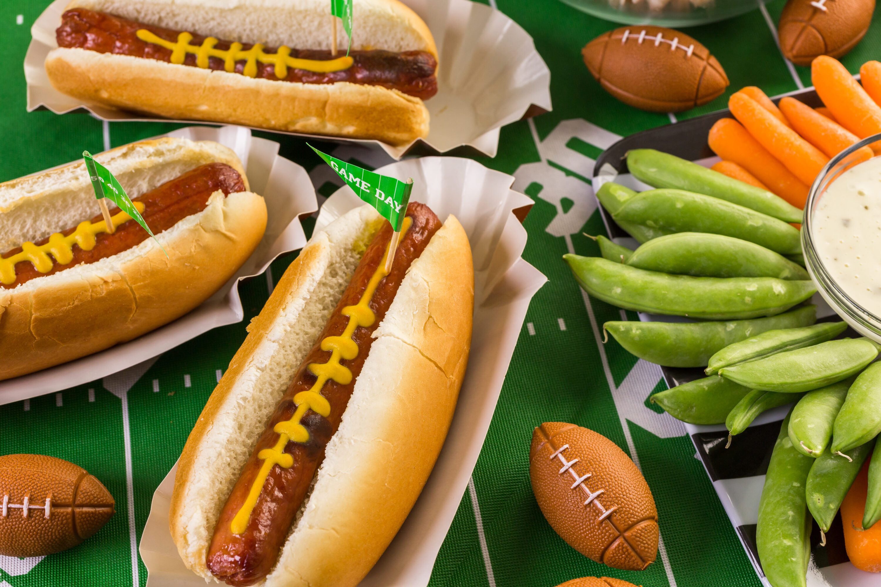Game day table with hotdogs, a veggie tray, and miniature footballs.
