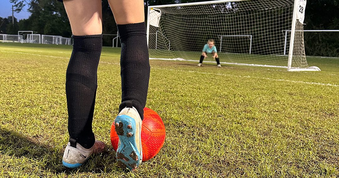 zoom in on soccer cleats about to kick a red soccer ball to the goal where a child goalie stands. image used to represent young athletes with type 1 diabetes