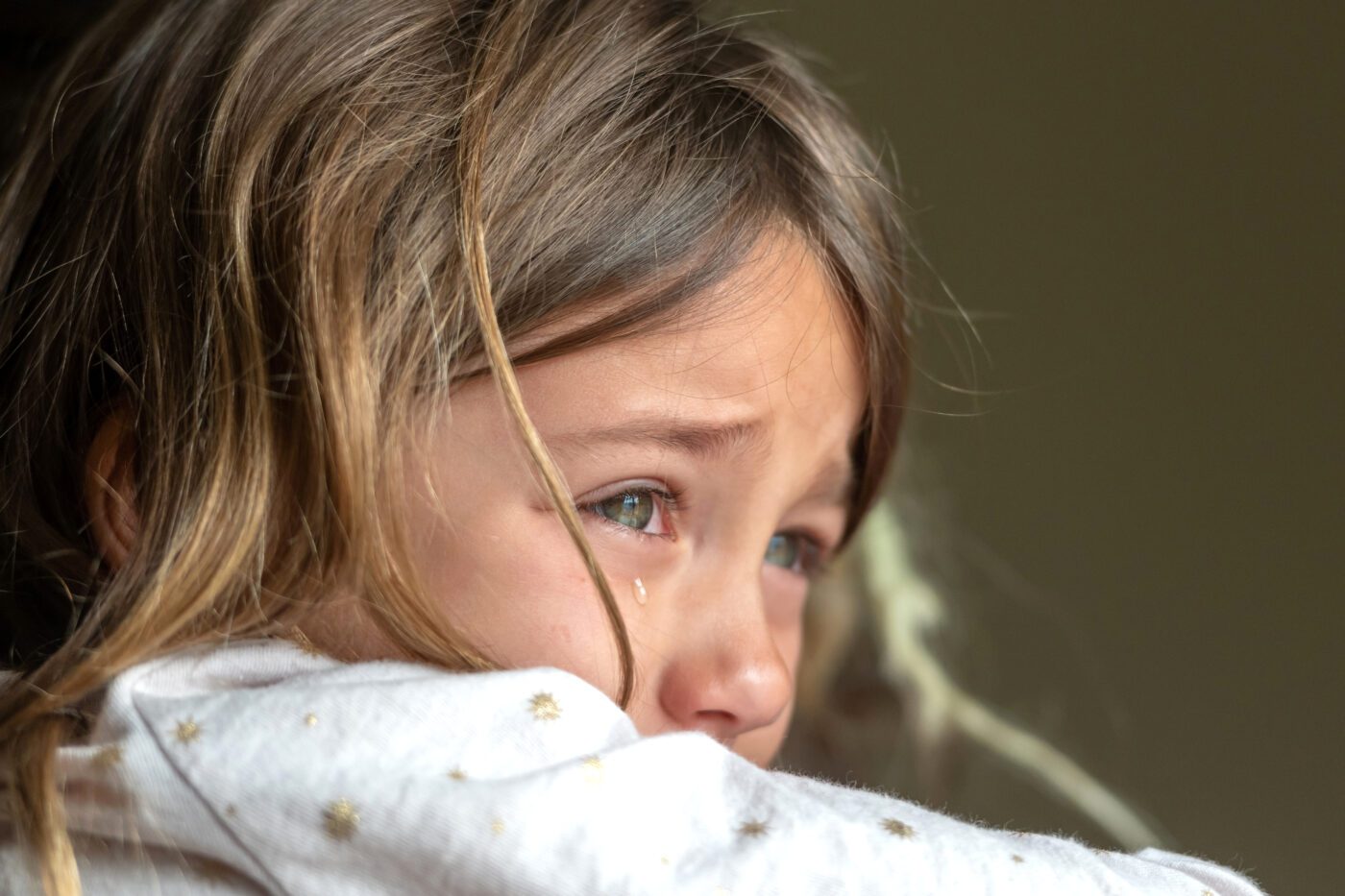 Close-up of a child with a tear on their cheek, resting their face against a sleeve and looking to the side.
