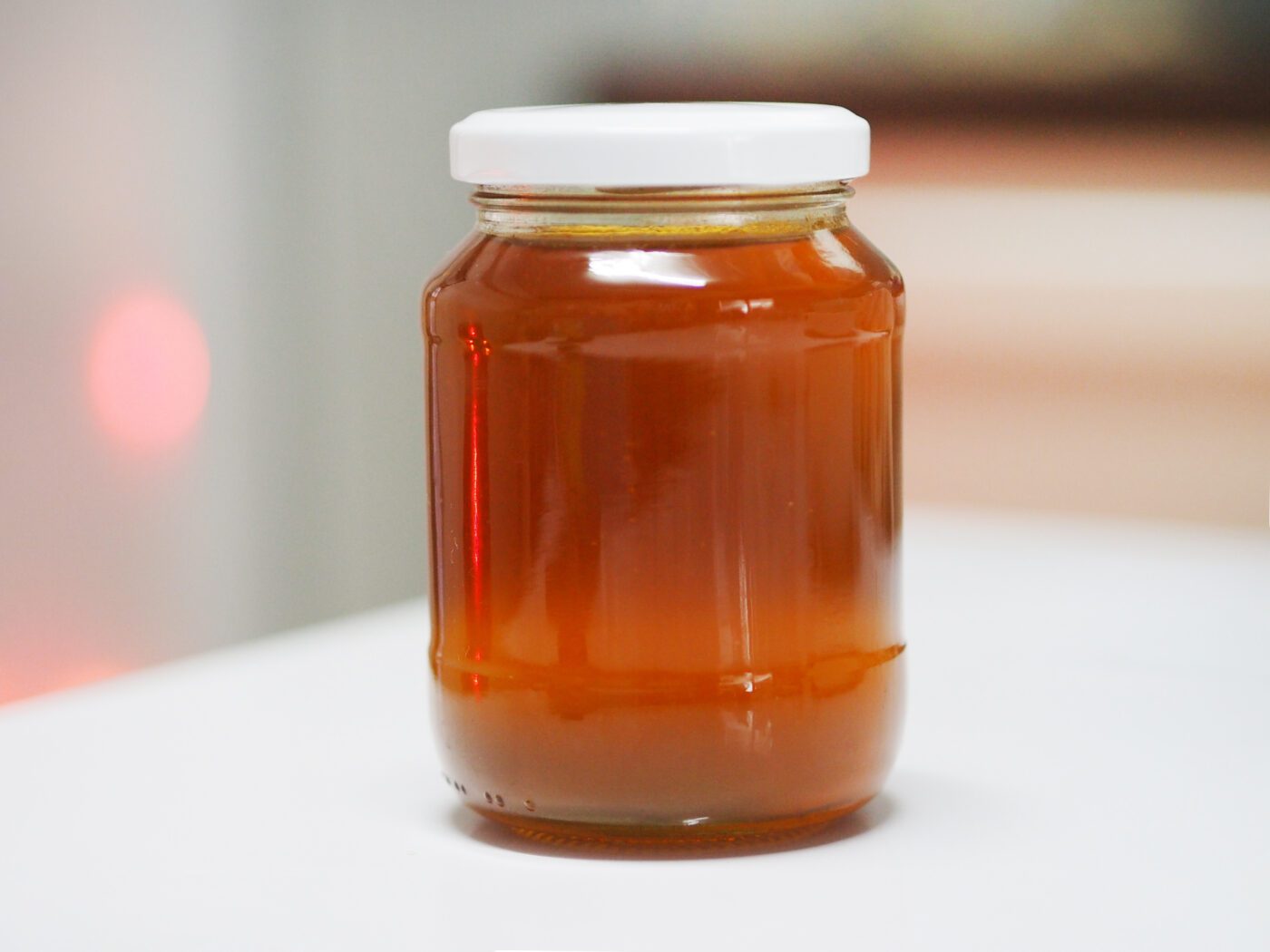 A clear glass jar filled with amber-colored honey and sealed with a white lid sits on a white surface.