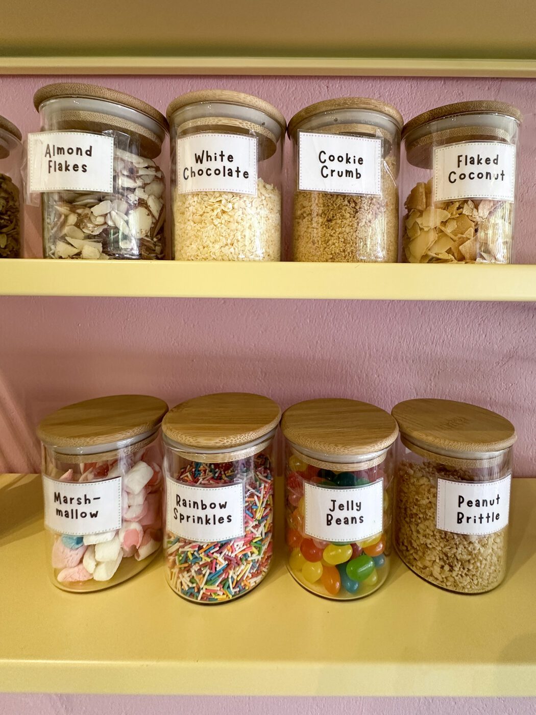 Glass jars on shelves filled with baking ingredients including almonds, white chocolate, cookie crumbs, coconut, marshmallows, sprinkles, jelly beans, and peanut brittle.