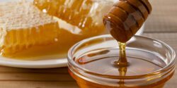 Honey dripping from wooden dipper into a glass bowl with honeycomb on a plate in the background.