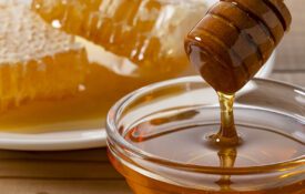 Honey dripping from wooden dipper into a glass bowl with honeycomb on a plate in the background.
