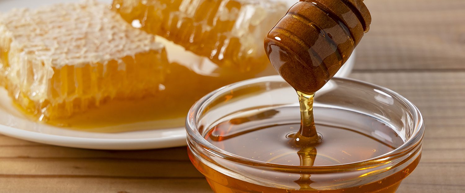 Honey dripping from wooden dipper into a glass bowl with honeycomb on a plate in the background.
