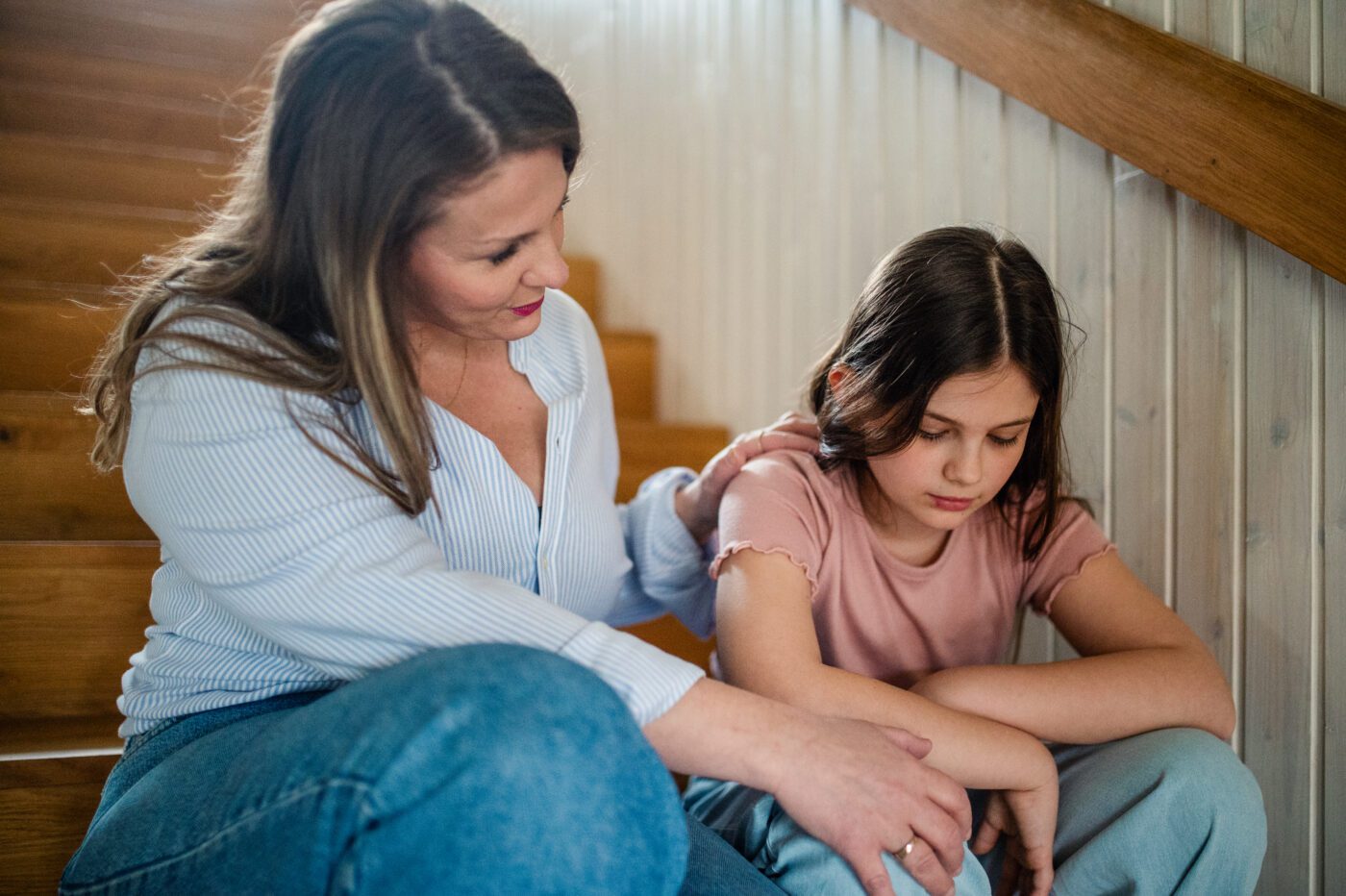 A woman sits beside a girl on wooden stairs, resting one hand on her shoulder and the other on her forearm. The girl looks down with her arms folded across her knees.