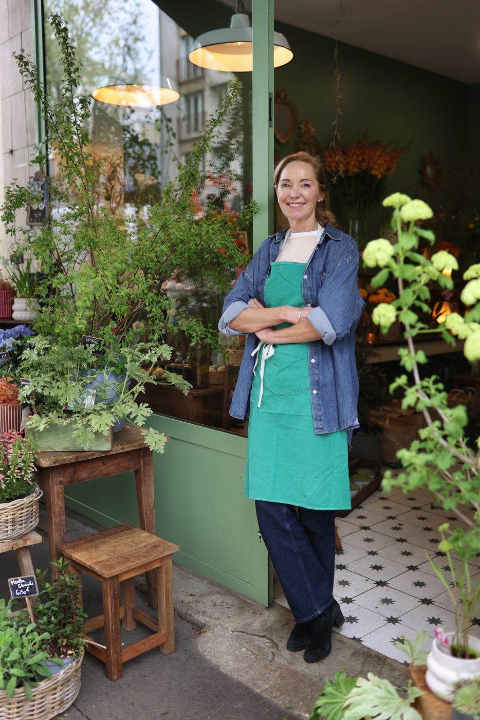 A smiling woman wearing a green apron and denim shirt stands with her arms crossed in the doorway of a flower shop, surrounded by potted plants and flowers.