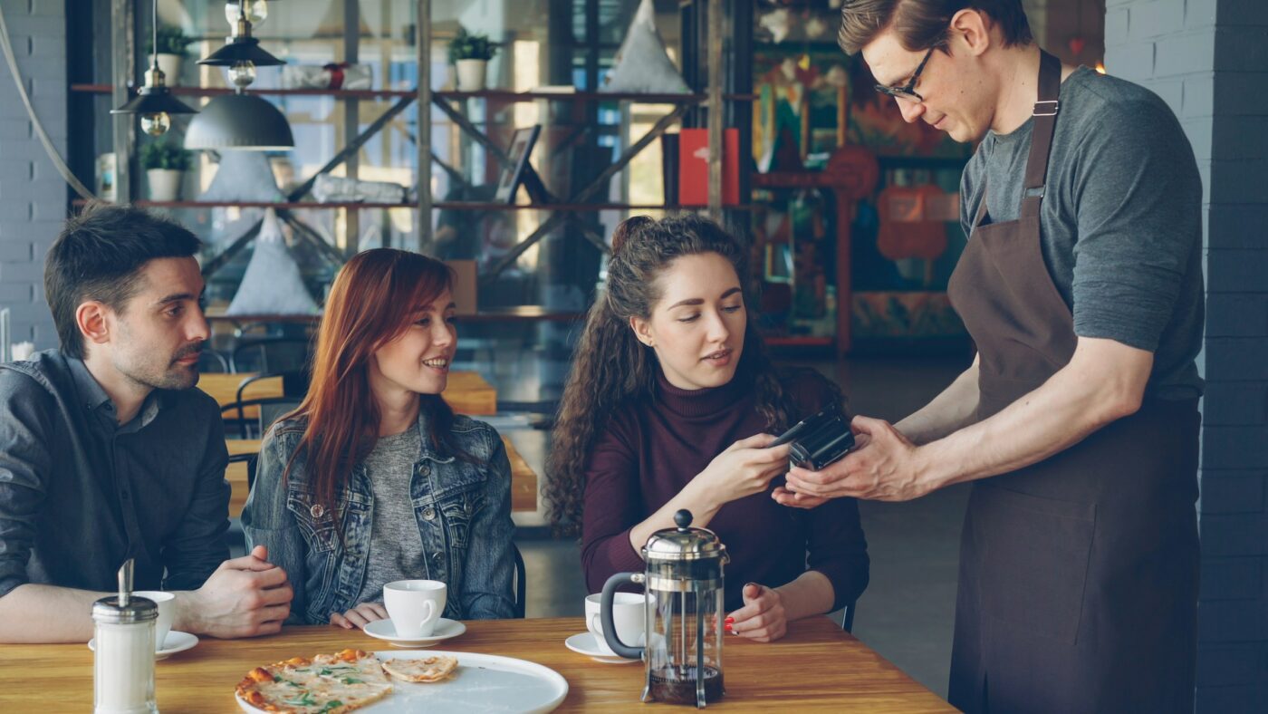 Two young women and a young man sitting at a pizza restaurant table with one of them paying a male waiter.